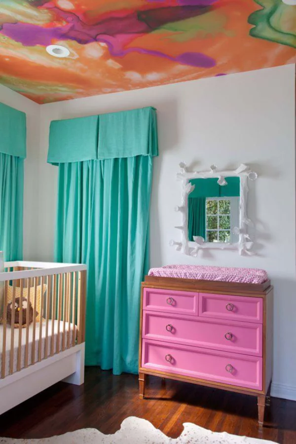 Kunterbuntes Babyzimmer bunt gestrichene Zimmerdecke türkisblaue Gardinen weißes Bett rosa Wickeltisch